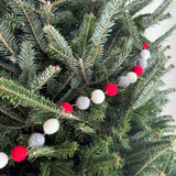 A felt festive pompom ball garland is draped on a lush, green Christmas tree. The garland features alternating red, gray, and white felt balls, adding a warm, celebratory touch to the festive branches.