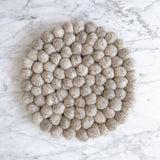 An 8-inch gray felt ball round pot trivet rests on a bright marble surface. This handmade felt coaster is crafted from numerous hand-stitched 100% organic lamb wool felt balls, each about an inch. Its textured surface and cool gray tones create a simple yet refined, eco-friendly accent for any home.