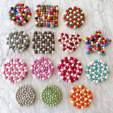 A gray felt ball round placemat is prominently displayed among other felt ball placemats on a bright marble surface. This handcrafted felt placemat, ethically made by Nepalese female artisans, is composed of tightly packed, hand-stitched gray felt balls, each about an inch in size. Other customizable felt pieces in various shapes and colors are also shown, highlighting the use of 100% organic lamb wool and azo-free dyes.