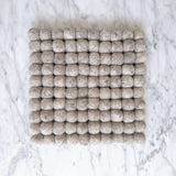 An 8-inch gray felt ball square pot trivet or placemat rests flat on a white marble surface. This handcrafted trivet, made from ethically sourced organic wool felt, features numerous small felt balls meticulously hand-stitched together to form a uniform square shape. Its cozy texture and neutral gray hue provide a calming, natural look, highlighting its fair-trade craftsmanship by Nepalese female artisans.