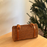 A woven rattan handbag, brown in color with visible texture, rests on a wooden surface, likely a table or desk. In the background, a Christmas tree is visible, suggesting a holiday setting. The bag features an adjustable leather handle and Batik lining, highlighting its eco-friendly nature and unique design elements.