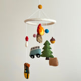 A camping wool felt baby mobile is displayed against a bright white backdrop. This nursery baby mobile features a circular white felt frame from which various felt figures suspend. These include a blue minivan, a green tree, an orange and yellow campfire, and a brown bear-adorned kayak. Small yellow, pink, and blue felt balls are also above the frame, all crafted with soft textures for a cozy, whimsical scene.