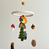 A Camping wool felt baby mobile is displayed against a plain, light background, featuring handcrafted elements. From a white felt ring, various natural New Zealand sheep wool felt figures are suspended, including an orange and yellow campfire, a green pine tree, a blue minivan, an acorn, a red and white mushroom, a classic tent, a bear-adorned kayak, and a brown backpack. The soft textures create a gentle, comforting nursery atmosphere.