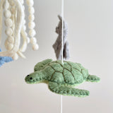 This handcrafted wool felt baby mobile, designed with a calming ocean theme, hangs from a wooden frame against a plain white background. A vibrant green sea turtle with a textured shell and dark green stitching is prominent, while a gray felt dolphin and a white jellyfish with hanging felt balls and tentacle-like strands are visible, creating a delightful nursery display.