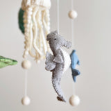 A charming wool felt baby mobile featuring sea life creatures is displayed, centered within a visible wooden frame. At its heart is a light gray seahorse with a white stomach and dark gray fins. Surrounding it are a white jellyfish with a cap and flowing tendrils above, a green felt turtle to the left, and a blue felt dolphin to the right. Small white spheres suspended by cotton threads enhance the calm, enchanting scene.