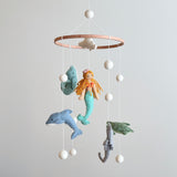A charming wool felt baby mobile, titled 'Mermaid Baby Mobile', hangs from the ceiling, featuring a mermaid at its center surrounded by sea turtles, sea horses, whales, dolphins, and stingrays. The mobile is suspended from a wooden frame, adding rustic charm to the scene.