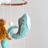 A felt sea life baby mobile, featuring mermaids, sea turtles, sea horses, whales, dolphins, and stingrays, hangs from a wooden hanger. The soft pastel colors evoke a sense of tranquility. Made from 100% wool felt, this handmade mobile is crafted with love in Nepal.