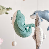 A close-up of a wool felt Sea Life baby mobile features several suspended sea creatures against a plain white background, creating a serene atmosphere. A light-green whale with a white belly hangs centrally, with a gray sea horse to its right. Above, a blue dolphin and a partially visible light-green sea turtle complete the tranquil ocean theme.