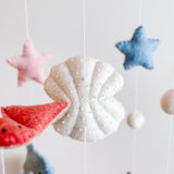 A wool felt Sea Life Baby Mobile is shown in a close-up against a plain white background. Various wool felt sea creatures, including scallops, sea turtles, seahorses, whales, dolphins, and stingrays, hang from a wooden frame by cotton threads. An off-white beaded scallop and a red sea creature are visible, creating a serene marine theme ideal for a nursery.