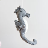 A gray wool felt seahorse, part of a sea life baby mobile, hangs centrally from a thin white string within a wooden frame against a plain white background. The seahorse features a distinctive curled tail and a white belly, with a visibly soft, plush felt texture.