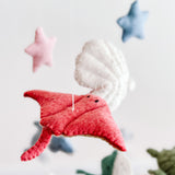 A red wool felt stingray, part of a whimsical sea life baby mobile, is prominently featured against a soft, off-white background. Crafted from textured red felt, the stingray is suspended by a thin white cotton thread and features small black button eyes. A visible wooden frame and partially seen wool felt scallop complete the playful, serene oceanic scene.