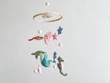 A Sea Life wool felt baby mobile is displayed against a plain white background, creating a calm atmosphere. It features a wooden ring from which a green sea turtle, a teal whale, a blue dolphin, and a grey seahorse are suspended by thin white cotton threads. White wool felt balls are positioned at the top, in the center of the ring, and at the bottom of some threads, adding a playful touch perfect for a nursery.