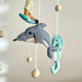 A delightful Surfer Boy and Ocean Creatures felt baby mobile hangs in a close-up studio shot against a soft beige background. This serene mobile showcases a gray felt dolphin with a cream underbelly and stitched features, a light turquoise felt seahorse with a curved tail, and a surfer riding a turquoise board. All components are suspended by white strings accented with light beige wooden beads, creating a whimsical ocean scene.