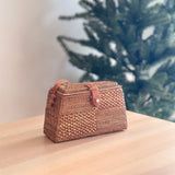 A Bali Rattan Crossbody Bag, titled 'Maya', rests on a wooden surface, showcasing its rattan material with a basket weave pattern. The bag features a rectangular shape with rounded corners, a flap closure secured by a leather snap button, and an adjustable strap for versatile wear. This stylish accessory complements the festive setting of a Christmas tree in the background.