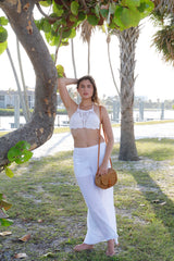 A person stands outdoors, surrounded by palm trees, wearing a white crop top and high-waisted pants, along with a straw hat. They lean against a tree, holding the Bali Rattan Crossbody Bag - Guitar, made of rattan material with an adjustable strap and snap closure.
