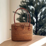 A woven rattan handbag, brown with visible weaving patterns, sits on a table amidst a festive Christmas tree setting. The bag features a sturdy handle made of natural material and has a secure leather hook closure. Its oval shape and unique Batik cotton lining add to its elegant and durable design.