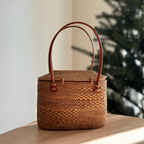 A woven rattan handbag rests on a surface, positioned in front of a softly blurred Christmas tree. The bag features a smooth finish, handle, and hook-and-loop closure mechanism. A geometric pattern is visible on the bag's surface, evoking a sense of holiday cheer.