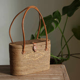 A handwoven basket-style rattan handbag, made from natural rattan material in Bali, rests on a wooden surface. The rattan shoulder handbag features leather handles, a lid with hook closure, and a medium size suitable for carrying essentials.