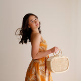 A woman stands against a white background, holding a woven rattan handbag with a batik lining and drawstring pouch. The bag is made from natural materials, possibly Ata grass, with a textured surface. It's spacious for daily essentials and features a unique design.