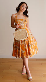 A woman stands against a plain background, wearing a bright orange dress with a floral pattern and styled hair. She holds a small, woven purse made from natural materials like rattan or similar, featuring a textured surface and drawstring closure.