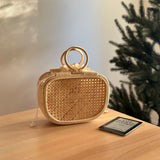 A wicker handbag, Charleston style from Bali Rattan Handbag, rests on a wooden table next to a smartphone. The bag's textured surface and visible handle are showcased against the cozy background of a Christmas tree and a wall with a framed picture. The handbag features a batik lining drawstring pouch for added functionality.
