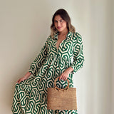 A woman stands in front of a white wall, wearing a green and white patterned jumpsuit with a belted waist. She holds a woven basket made from rattan or natural material, posing with one hand on her hip. The product, Bali Rattan Handbag - Kelsey, is the focus of the image.