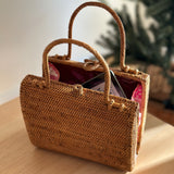 A woven rattan handbag rests on a wooden table, showcasing its natural brown color and traditional Balinese Batik pattern. The bag features a sturdy base for upright placement and short handles made from Ata grass.