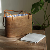 A detailed, eye-level shot of a rectangular, handwoven rattan tote bag resting on a rustic wooden table. The bag, golden-brown in the natural sunlight, features thin, tan leather shoulder straps attached to its rim. The open top reveals a crisp, blue and white vertically striped fabric lining. In the foreground, a silver tablet with a folded white cover lies on the table. To the right, the blurred, deep green leaves of a monstera plant create a natural backdrop, while soft light from the left casts a gentle