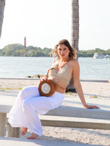A woman sits on a bench by the water, wearing a white top and holding a woven straw hat. In front of her is a wooden table with a small plant, set against a serene body of water with a boat in the distance. The scene evokes a relaxed, leisurely atmosphere, suggesting a vacation or beach day setting. A Bali Round Rattan Crossbody Bag, adorned with real seashells, is implied by the context.