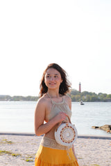 A young woman stands on a sandy beach near the water's edge, holding a beige crossbody bag with intricate seashell detailing crafted from rattan material. The background features a clear sky and calm waters, evoking a peaceful atmosphere.