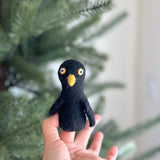 A black raven felt finger puppet is presented by a female hand, set against a blurred Christmas tree background. This unique handmade felt finger puppet is crafted from black felt, featuring prominent wings and a yellow felt beak. It has distinctive yellow and white eyes, highlighting its handcrafted detail.
