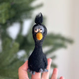 A handmade wool felt black sloppy bird finger puppet is held up by a female hand, against a soft, out-of-focus background with a hint of a Christmas tree. This unique felt finger puppet features a black body, a fluffy yellow beak, and expressive white eyes with gray accents. Gray wool forms a tuft on its head and its feet, giving it a playful, festive charm.