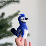 A handcrafted wool felt blue jay finger puppet, made by fair-trade artisans, is gently presented by a female hand against a soft, naturally lit background with a hint of a Christmas tree. This vibrant blue jay felt finger puppet features striking blue feathers on its head, wings, and tail, a distinctive crest, a white belly, and black accents around its beak, highlighting its realistic charm.