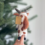 A handmade wool felt brown cow finger puppet is held upright by a female hand, set against a softly blurred Christmas tree background. This unique felted finger puppet features distinct brown and white spots, small horns, and a small tag attached, reflecting its detailed craftsmanship.