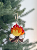 A cozy Campfire with marshmallow felt ornament hangs on a vibrant green Christmas tree branch. This handmade felt ornament features layered red, orange, and yellow flames rising from brown felt logs detailed with lighter wood grain. Three white felted marshmallows are roasted atop the fire, and a jute string allows for easy hanging.