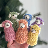 Six pastel felt sheep finger puppets, known as 'Candy Sheep', are held in a hand against a soft, natural backdrop featuring a Christmas tree. Each handmade felt sheep finger puppet, colored pink, green, orange, blue, purple, or yellow, boasts a soft, fluffy 3D design. They feature small white felt ears and a simple, endearing embroidered expression, perfect for imaginative play.