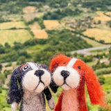 An orange needle felted Cavalier King Charles Spaniel finger puppet stands outdoors amidst green fields and trees. This handcrafted felt kids toy features a soft texture with long, fluffy, floppy ears and loving eyes, creating a heartwarming scene.