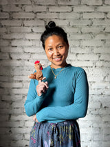 A light brown handmade felt chicken finger puppet is prominently displayed on the finger of a smiling Nepalese female artisan, against a soft white brick wall. This unique wool felt chicken finger puppet features a distinctive red crest and beak, showcasing its intricate handcrafted details. This charming felt toy for kids highlights the skilled artistry and fair-trade craftsmanship of artisans in Nepal.