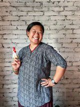 A handcrafted wool felt chicken finger puppet, expertly made by a Nepalian artisan, is held up against a textured white brick wall. This unique felt chicken finger puppet, designed for engaging finger play, is primarily white with a vibrant red comb and a yellow beak, small enough to fit on a fingertip.