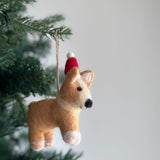 A charming felt corgi ornament, also a festive Christmas dog ornament, dangles from a green Christmas tree branch against a soft white backdrop. Crafted from soft, light brown felt, this corgi wears a red Christmas hat with a white pompom. It has small upright ears, a tiny black nose, and white markings on its paws, adding a heartwarming touch to holiday decor.