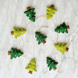 A felt Christmas Tree Garland with jute thread is displayed on a mottled white surface. It features alternating green and light green felt Christmas trees. Each tree is embellished with small white embroidered snowflakes and has a brown felt tree trunk, connected by a thin jute thread for natural texture.