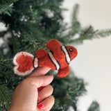A handcrafted wool felt clown fish finger puppet is presented held by a female hand against a blurred green Christmas tree. This vibrant orange clown fish features googly eyes and distinct white stripes outlined in black, making it a delightful felt toy for kids. Made from soft sheep wool, this unique finger puppet adds a cheerful touch for imaginative play.