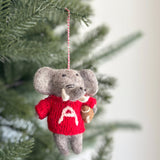 An Alabama elephant Christmas ornament hangs on a Christmas tree, radiating holiday cheer and team spirit. This handcrafted wool felt elephant ornament is gray, featuring expressive black eyes and a cheerful smile. It wears a bright red sweater with a white 'A' and holds a brown football, perfect for college football fans. A red and white string is attached for hanging.