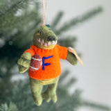 A green wool felt Florida gator Christmas tree ornament hangs on a green Christmas tree. The handcrafted felt gator wears a bright orange shirt with a blue 'F' and holds a brown football with white stripes. A jute string is attached for hanging, adding a festive touch to the holiday season.