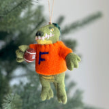 A handcrafted wool felt Florida Gator Christmas ornament, designed for team spirit, hangs on a festive Christmas tree. This playful green gator wears an orange sweater with a blue 'F' and holds a football, with yellow and pink details around its mouth. A jute string is attached for easy hanging.