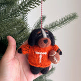 A unique wool felt Tennessee Volunteers dog ornament from the College Football Collection is gently held by a female hand, suspended from a pine branch against an off-white backdrop. This handmade felt ornament features a black dog with a white head stripe, brown muzzle, and ears. It proudly wears an orange sweater with a white 'T' and clutches a football, showcasing detailed hand-stitching and a red and white hanging string.