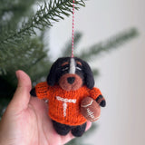 A female hand gently holds a Tennessee Volunteers wool felt dog ornament, suspended by a red and white string from a blurred Christmas tree. This charming black and brown dog ornament wears an orange sweater with a white 'T' and clutches a football, featuring hand-stitched details for an artisanal feel.