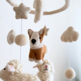A wool felt Corgi and Sheep Baby Mobile hangs against a bright white background, showcasing a whimsical, neutral-colored nursery decor item. A smiling brown, white, and black wool felt corgi is central, surrounded by fluffy white wool felt sheep, stars, clouds, and felt balls. All elements are suspended from a circular wooden frame, creating a cozy farmhouse aesthetic.