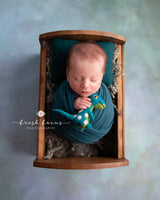 A handcrafted Brontosaurus wool felt finger puppet is gently cradled in a baby's hands, nestled in a rustic wood bed with a soft gray blanket. This unique, needle-felted dinosaur features a lifelike 3D design with a long neck, blue-green body, white spots, and a yellow eye. Handmade from 100% wool, this fair-trade puppet adds a comforting element for imaginative play.