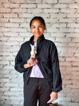 A light gray needle-felted dire wolf finger puppet is held on a smiling female model's finger, showcased against a white brick wall. This handcrafted felt toy is made from soft wool and features a small black nose, embodying the mythical dire wolf's spirit.